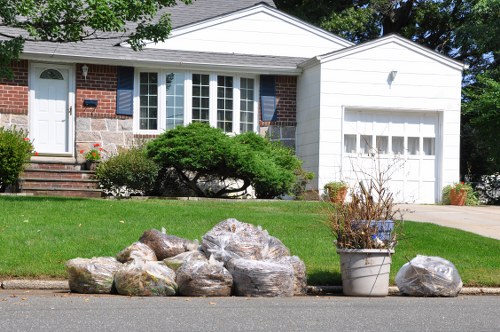 In front of a residential property in Woolwich, a collection of large, irregularly shaped rocks and stones in shades of grey and brown are neatly arranged on the pavement beside a green lawn. To the right, a white plastic bucket contains dried, leafless branches or plant debris. The background features the front of a single-story house with a brick exterior, large white framed windows, and a white garage door. The lawn is well-maintained, and a small bush with dense green foliage partially obscures the house's front steps. The scene appears to be part of garden or yard clearance, with Garden Clearance Woolwich potentially involved in removing waste or debris as part of garden maintenance or rubbish removal services in the area. The overall setting is bright and clear, with natural sunlight illuminating the outdoor scene.