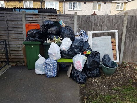 A collection of black, white, and patterned rubbish bags filled with various waste materials is piled on and around a green waste bin and a plastic outdoor chair in a small backyard area. The debris includes plastic bags, some transparent, revealing mixed waste inside, and others opaque. The scene is set against a wooden fence with horizontal panels, behind which part of residential buildings with mosaic-style windows are visible. To the right of the rubbish, a large, empty portable frame and a plastic tub are positioned on bare soil. The ground in the foreground is paved with concrete, and the overall environment appears to be a typical urban garden space in Woolwich, showing evidence of a recent rubbish clearance service by Garden Clearance Woolwich, focusing on waste removal from residential gardens.