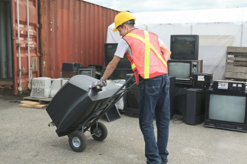 A man wearing a yellow safety helmet and a high-visibility vest with reflective strips is pushing a black outdoor wheeled trash bin. He is in an outdoor area that appears to be a waste collection or disposal site, with a row of old, used electronic appliances such as televisions, radios, and speakers stacked against a corrugated metal wall. The ground is paved with asphalt, and there is a wooden pallet nearby on the left side of the image. In the background, there is a translucent white roof covering part of the area, providing shelter for the stored items. The scene is well-lit, likely under natural daylight, and the environment suggests an area used for rubbish collection or clearance, consistent with services offered by Garden Clearance Woolwich in the local area around Woolwich and surrounding postcode regions.