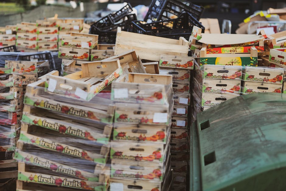 A large outdoor collection of empty wooden fruit crates, primarily stacked in multiple piles on a paved surface. The crates are light-colored wood with a smooth finish, featuring printed labels with i